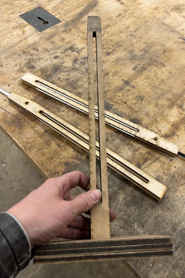 Holding up the assembled frame, 2 identical uprights slotted through a three-layer base. The uprights also have a vertical slot for adjusting the arms up and down. The assembled arms with pointer ends are on the table behind the frame.
